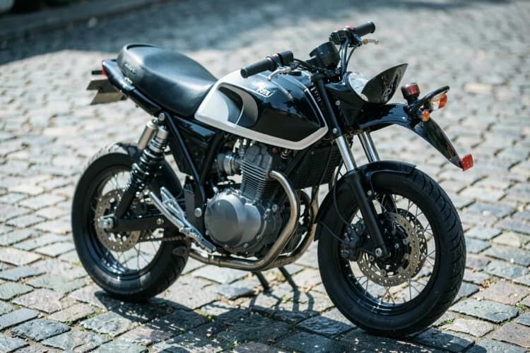 Uma moto Honda Fan 160 preta e branca estacionada em uma rua de paralelepípedos com luz solar.